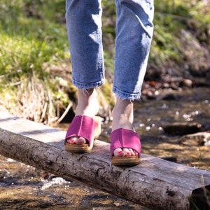 May include: A person wearing blue jeans and pink suede clogs with wooden soles walks across a wooden log over a stream.
