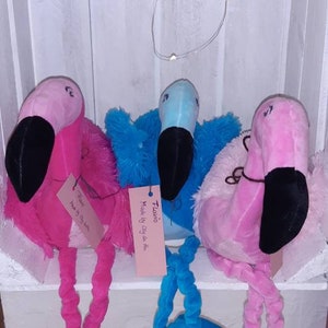 May include: Three plush flamingo toys with long legs. The flamingo on the left is pink with a white head and a tag that says "Flavo, Made by Day Lee Mia". The flamingo in the middle is blue with a white head and a tag that says "Flavo, Made by Day Lee Mia". The flamingo on the right is pink with a white head and a tag that says "Flavo, Made by Day Lee Mia".