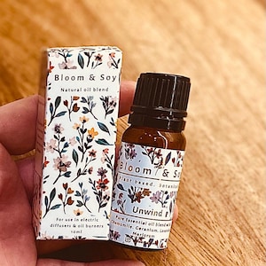 May include: A small brown glass bottle with a black lid and a white label that says "Bloom & Soy Plant based. Botanical Unwind Pure Essential oil blend with Chamomile. Geranium. Lavender. Marjoram" and a floral pattern. The bottle is in a hand holding a small white box with a floral pattern and the text "Bloom & Soy Natural oil blend For use in electric diffusers & oil burners 10ml".