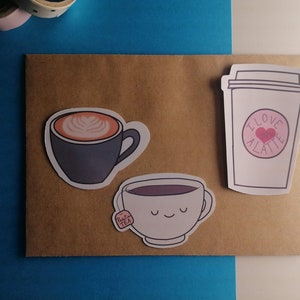 Op de afbeelding: Drie koffie- en theestickers op een bruine papieren achtergrond. De stickers zijn van een latte, een kop koffie met latte art en een theekopje met een theezakje.