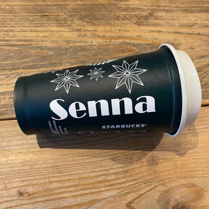 Op de afbeelding: Een donkergroene Starbucks koffiebeker met de naam "Senna" in witte letters. De beker heeft witte sterontwerpen en een wit deksel. Het Starbucks-logo is ook zichtbaar.