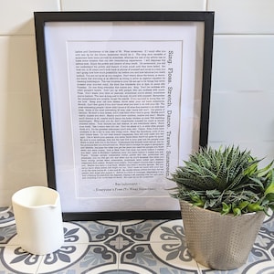 May include: A framed black and white print with a motivational quote about life advice. The quote is by Baz Luhrmann and is from the movie "Everyone's Free to Wear Sunscreen". The print is sitting on a tiled surface with a white jug and a silver pot with a green succulent plant.