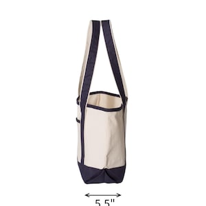 May include: Side view of a canvas tote bag with a cream-colored body and navy blue accents. The bag has navy blue handles and a navy blue bottom. The bag is 5.5 inches wide.