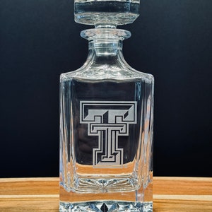 May include: A clear glass decanter with a square body and a stopper. The decanter has a  etched design of a stylized "T" on the front.