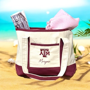 May include: A canvas tote bag with a maroon bottom and pocket, featuring the letters "A&M" and the name "Morgan". The bag is filled with a magazine and a pink waffle-weave towel. The bag has white handles and is on a sandy beach.