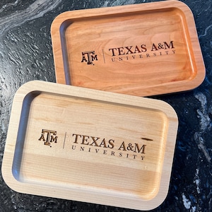 May include: Two rectangular wooden trays with rounded corners. One is light-colored, the other is a medium brown. Both trays feature the Texas A&M University logo and text etched into the wood. The trays are likely used for serving or display.