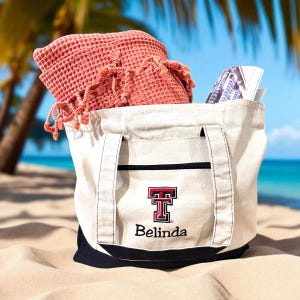 May include: A cream-colored canvas tote bag with a dark blue base and a front pocket. The bag features an embroidered red and black logo with the text "TT" and the name "Belinda" below it. A coral-colored waffle-weave towel with fringe is inside.