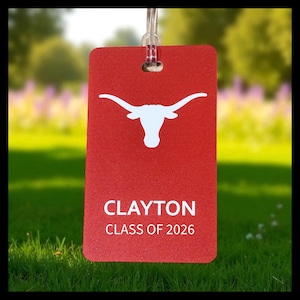 May include: A red luggage tag with a white longhorn logo and the name "CLAYTON" with "CLASS OF 2026" printed below. The tag is hanging from a clear plastic loop against a blurred green background.