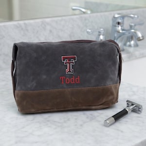 May include: A gray and brown canvas toiletry bag with the name "Todd" embroidered in red, along with a red and black "T" logo. A silver and black razor sits to the right of the bag. The bag is on a white marble surface.