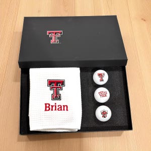 May include: A black gift box containing a white golf towel embroidered with "Brian" and a red and black logo, along with three golf balls. The golf balls have the same logo and "Texas Tech" printed on them.