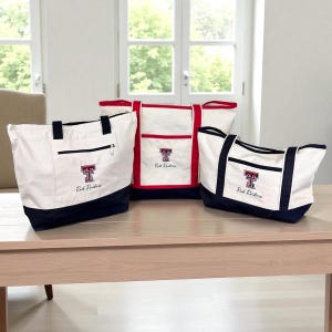May include: Three canvas tote bags in varying sizes, featuring a white body with navy blue accents and a red trim. Each bag displays the Texas Tech Red Raiders logo and text. The bags are on a light wood table.
