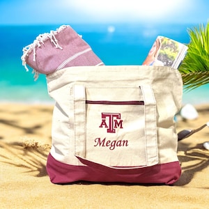 May include: A beige canvas tote bag with a maroon bottom and pocket, personalized with the name "Megan" and a maroon logo. The bag contains a folded pink towel and a magazine. The scene is set on a sandy beach with a blue ocean background.