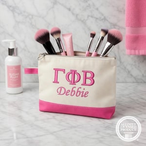 May include: A cream-colored makeup bag with a pink bottom and handle, embroidered with the Greek letters ΓΦΒ and the name "Debbie" in pink. The bag contains makeup brushes and a tube of lip gloss. A bottle of Sugar Plum lotion is to the left.