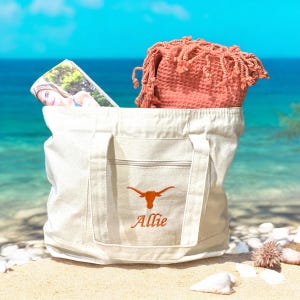 May include: A beige canvas tote bag with an orange longhorn logo and the name "Allie" embroidered on the front. The bag is filled with a coral-colored waffle-weave towel and a magazine. The scene is set on a sandy beach with a blue ocean in the background.