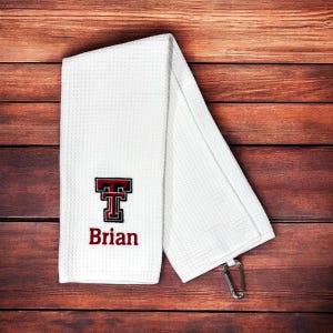 May include: A white waffle-weave golf towel featuring an embroidered red and black Texas Tech logo above the name Brian in red lettering. The towel includes a silver metal carabiner clip attached to the bottom corner for easy hanging.
