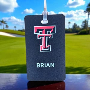 May include: A black luggage tag with the name "BRIAN" in white text. The tag features a red and white "T" logo. The tag is hanging against a backdrop of a golf course and blue sky.