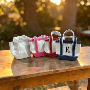 May include: Four small canvas tote bags in a row, each with a different color trim and handle. The bags are white with trim in gray, pink, red, and navy blue. The navy bag has a script letter "K" embroidered on the front.