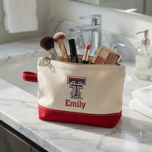May include: A canvas makeup bag with a red base and handle, and a cream-colored upper section. The bag features a red and black logo with the name "Emily" in red. The bag is open, revealing makeup brushes, mascara, lipstick, and an eyeshadow palette.