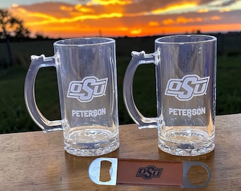 Könnte beinhalten: Zwei Bierkrüge aus klarem Glas mit Griffen, mit den Buchstaben "OSU" und dem Namen "Peterson" in Weiß. Ein brauner Lederflaschenöffner mit dem "OSU"-Logo ist ebenfalls vorhanden. Die Artikel befinden sich auf einer Holzoberfläche.