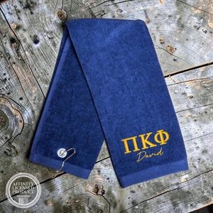 May include: A navy blue golf towel featuring a silver metal grommet and hook. The towel is embroidered with the gold Greek letters Pi Kappa Phi and the name David in a cursive script. The fabric has a soft, textured finish throughout the surface.