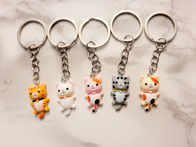 May include: Five colourful cat keyrings with silver key rings. Each keychain features a different cat design with orange, white, pink, grey, and cream colours.