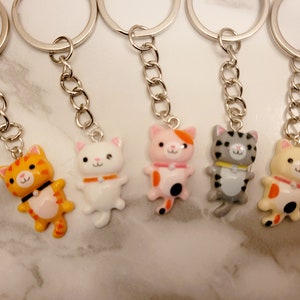 May include: Five colourful cat keyrings with silver key rings and chains. The cats are orange and white, white, pink and orange, grey and white, and yellow and orange.