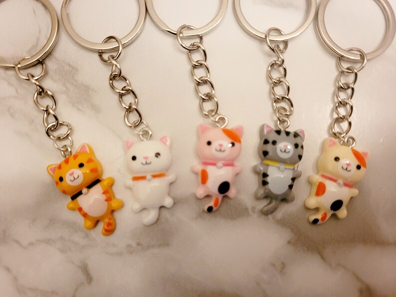 May include: Five colourful cat keyrings with silver key rings and chains. The cats are orange and white, white, pink and orange, grey and white, and yellow and orange.