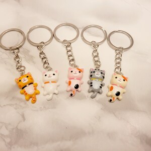 May include: Set of six keychains featuring different coloured cartoon cats with silver key rings. The cats are white, orange, pink, grey, and orange and white.
