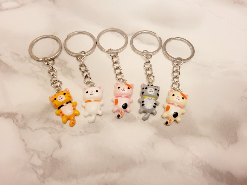 May include: Set of six keychains featuring different coloured cartoon cats with silver key rings. The cats are white, orange, pink, grey, and orange and white.