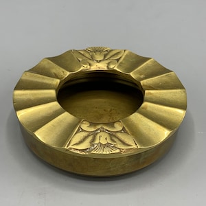May include: A round brass ashtray with a decorative design of stylized flowers around the rim. The ashtray has a fluted design on the sides.