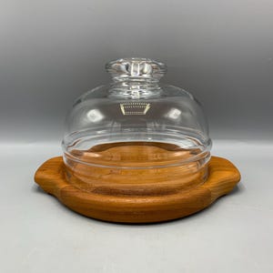Puede incluir: Una cúpula de vidrio transparente con una base de madera. La cúpula tiene un pomo en la parte superior y se asienta sobre una base redonda de madera con un borde festoneado.