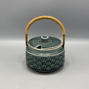 Puede incluir: Un azucarero de cerámica azul con un patrón floral y un asa de bambú tejido. La tapa tiene un pequeño pico para verter.