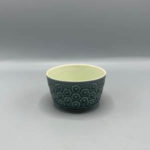 Puede incluir: Un pequeño cuenco de cerámica azul con un patrón floral. El cuenco tiene un interior verde claro y una base ligeramente elevada.