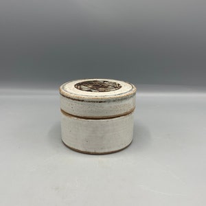 May include: A white ceramic jar with a brown geometric design on the lid. The jar is approximately 7.5 cm tall and 7.5 cm in diameter.