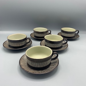 Puede incluir: Conjunto de cinco tazas de té y platillos de cerámica con rayas marrones y blancas. Las tazas y los platillos tienen un diseño simple y elegante.
