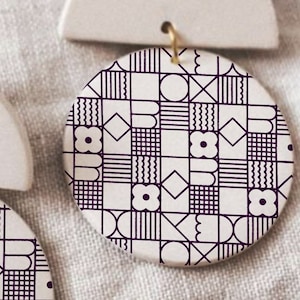 May include: A pair of white ceramic earrings with a geometric pattern in black. The earrings are shaped like a half-circle with a round disc attached to the bottom.