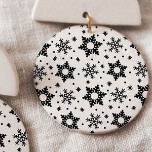 May include: A pair of white ceramic earrings with a snowflake pattern. The earrings are shaped like a half circle and a circle.