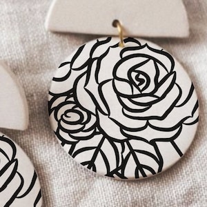 Peut inclure: Une paire de boucles d'oreilles en céramique blanche avec un design en demi-cercle et un pendentif rond orné d'un motif floral noir et blanc de roses.