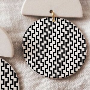 May include: A pair of white ceramic earrings with a black and white geometric pattern. The earrings are shaped like half circles with a round disc attached to the bottom.