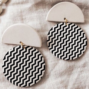 Pode incluir: Um par de brincos geométricos brancos e pretos. Cada brinco apresenta um semicírculo branco com um círculo de padrão chevron preto e branco anexado.