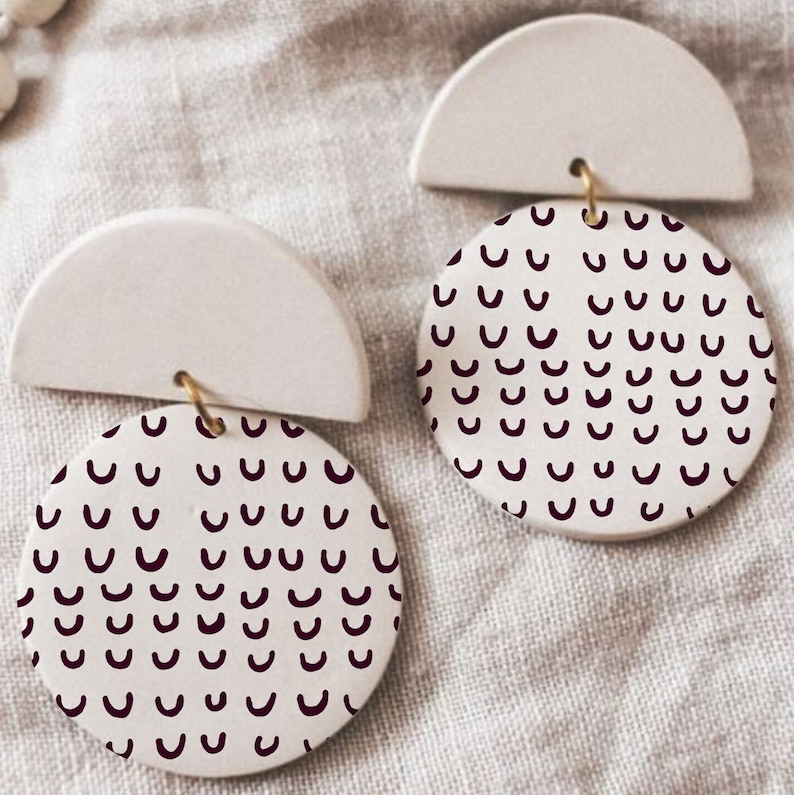 May include: A pair of white ceramic earrings with a half-circle design and a circular design featuring a repeating pattern of burgundy-coloured curved lines.