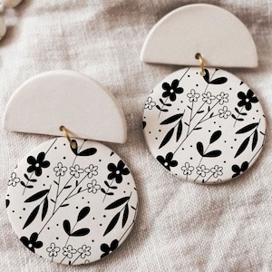 Może przedstawiać: Para białych ceramicznych kolczyków z czarno-białym wzorem kwiatowym. Kolczyki mają kształt półkola z okrągłym dyskiem przymocowanym do dołu.