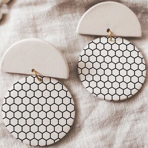 May include: A pair of white ceramic earrings with a black honeycomb pattern. The earrings are shaped like a half circle with a round disc attached to the bottom.