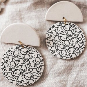 Può includere: Un paio di orecchini in ceramica bianca con un design a semicerchio e un pendente rotondo con un motivo di cerchi in bianco e nero.