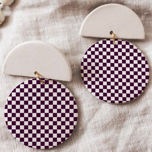 May include: A pair of white ceramic earrings with a checkerboard pattern in black and white. The earrings are shaped like a half circle with a round checkerboard pattern hanging from the bottom.