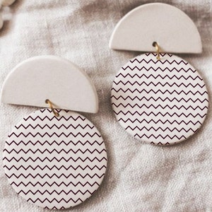 May include: A pair of white ceramic earrings with a burgundy chevron pattern. The earrings are shaped like a half circle with a round disc attached to the bottom.
