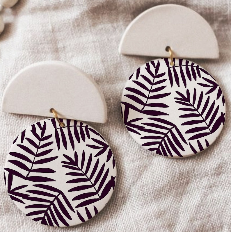 May include: A pair of white ceramic earrings with a black and white leaf pattern. The earrings are shaped like a half circle with a round pendant.