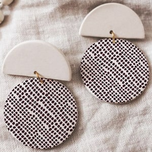 Könnte beinhalten: Ein Paar weiße Keramikohrringe mit einem Halbkreisdesign. Jeder Ohrring hat eine runde, burgunderfarbene Scheibe mit einem weißen Tupfenmuster.