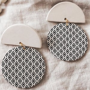 Peut inclure: Une paire de boucles d'oreilles en céramique blanche avec des cercles à motifs géométriques noirs et blancs. Les boucles d'oreilles ont une forme de demi-cercle sur le dessus et un cercle rond avec un motif sur le dessous.