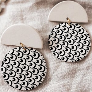 Può includere: Un paio di orecchini in ceramica bianca con un motivo geometrico bianco e nero. Gli orecchini hanno la forma di un semicerchio con un disco rotondo attaccato alla parte inferiore.